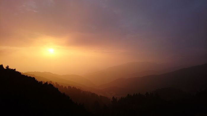 Спираща дъха фотография на силуета на пиринските гори на фона на залязващото слънце, което си проправя път измежду облаците и вечерната мараня
